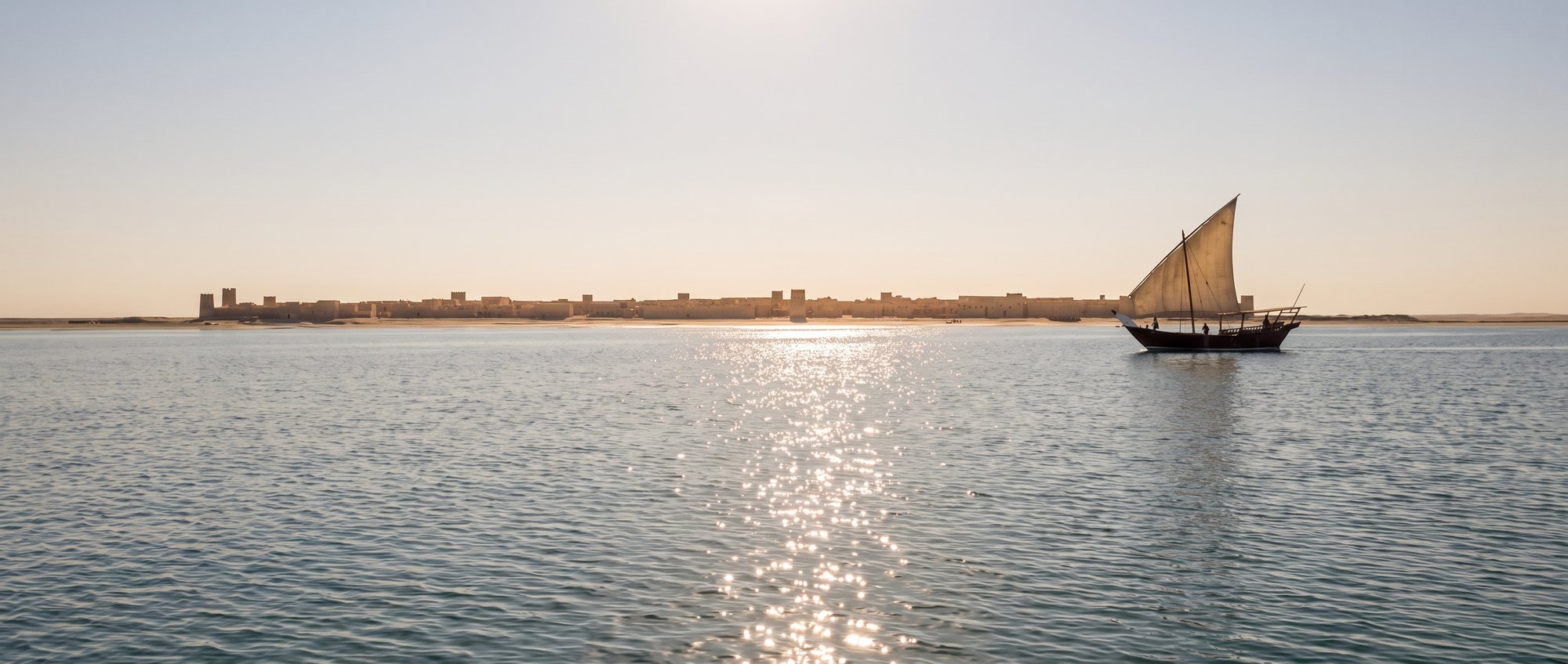 A dhow under sail on open water with the low skyline of Al Zubarah on the horizon